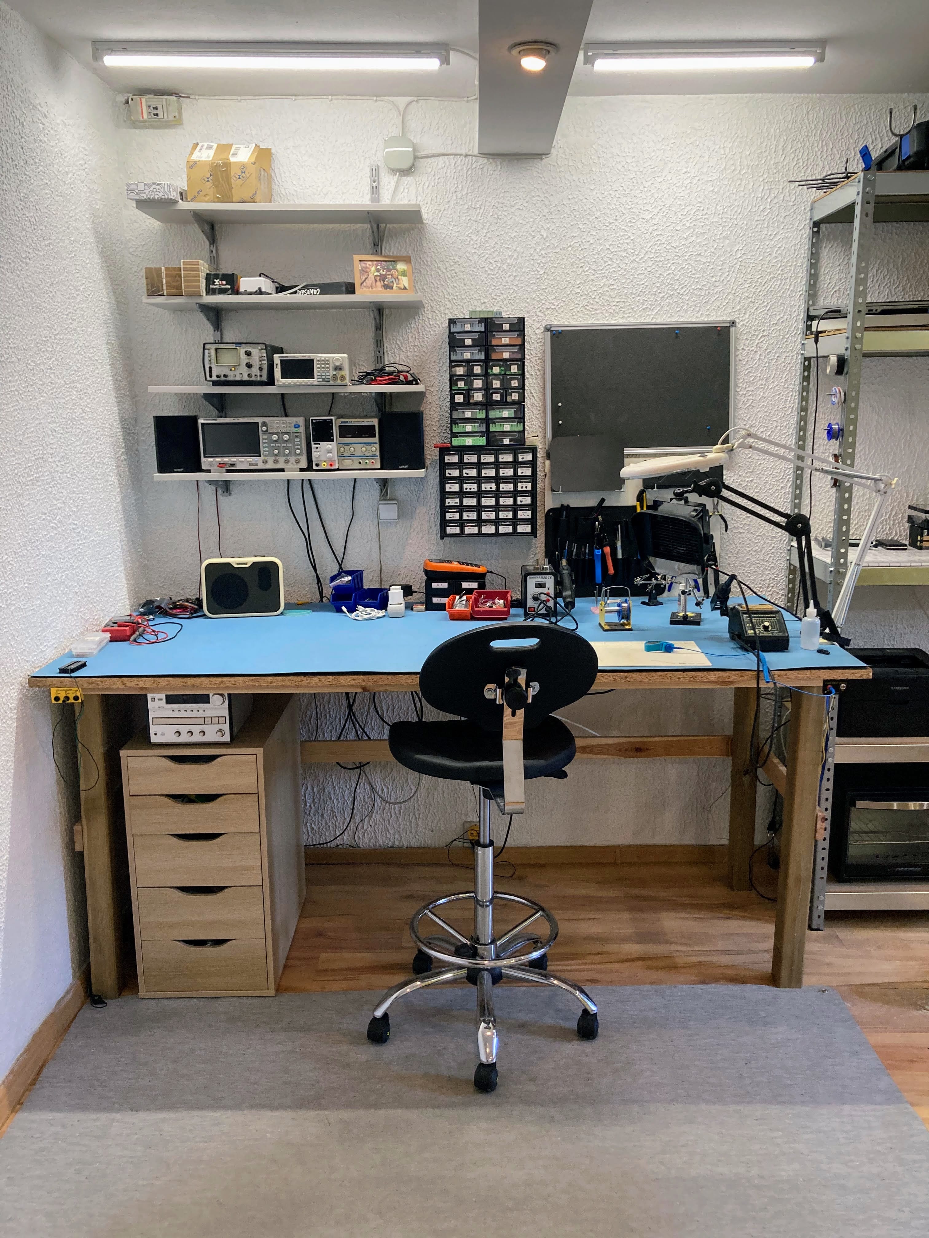 Zdjęcie warsztatu elektronicznego - stół warsztatowy z wysokim stołkiem i sprzętem laboratoryjnym na półkach.